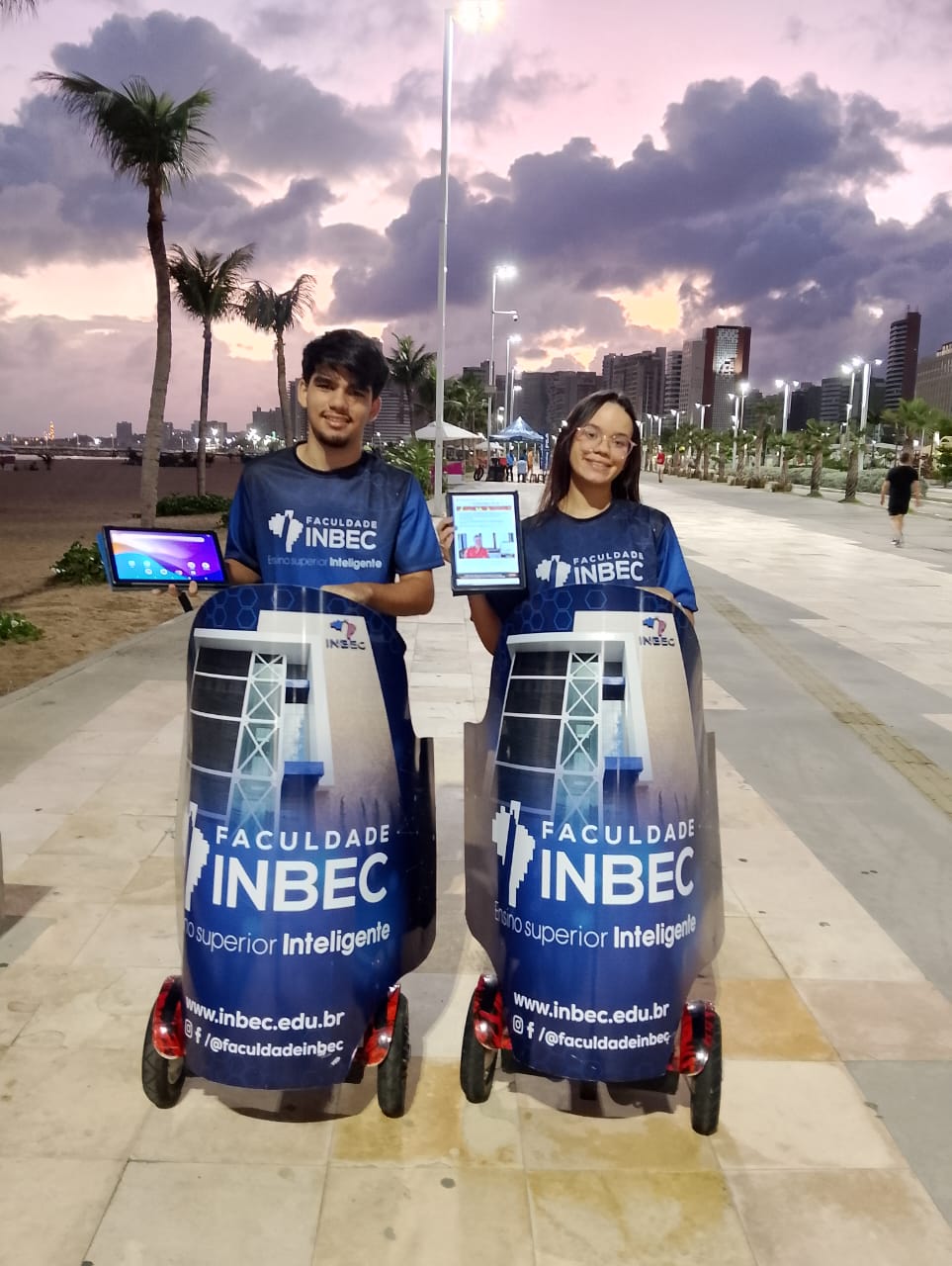 Ação de marketing da Faculdade INBEC na orla da Beira-Mar de Fortaleza. Dois promotores de evento utilizam segways adesivados com a marca e apresentam a faculdade em tablets para o público ao entardecer.