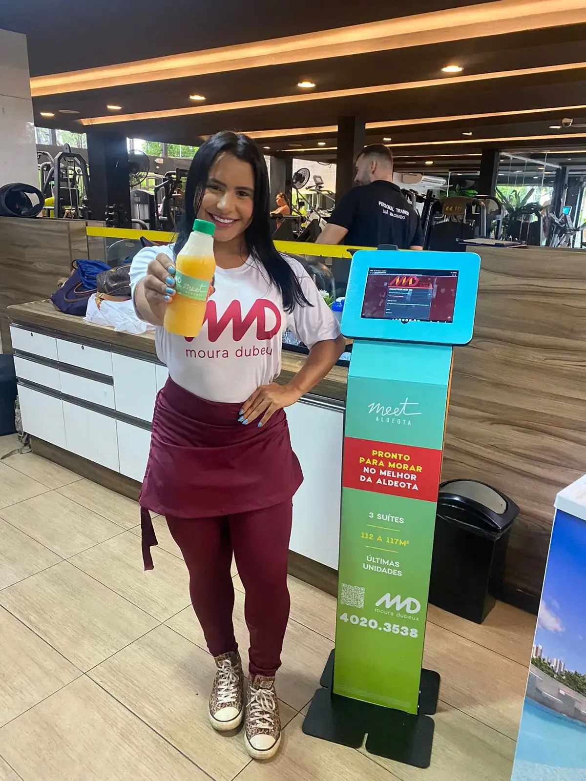Ativação de marca da construtora Moura Dubeux (MD) em uma academia para divulgar o empreendimento imobiliário Meet Aldeota. Uma promotora uniformizada distribui sucos como brinde para os frequentadores, ao lado de um totem digital interativo com informações do imóvel.