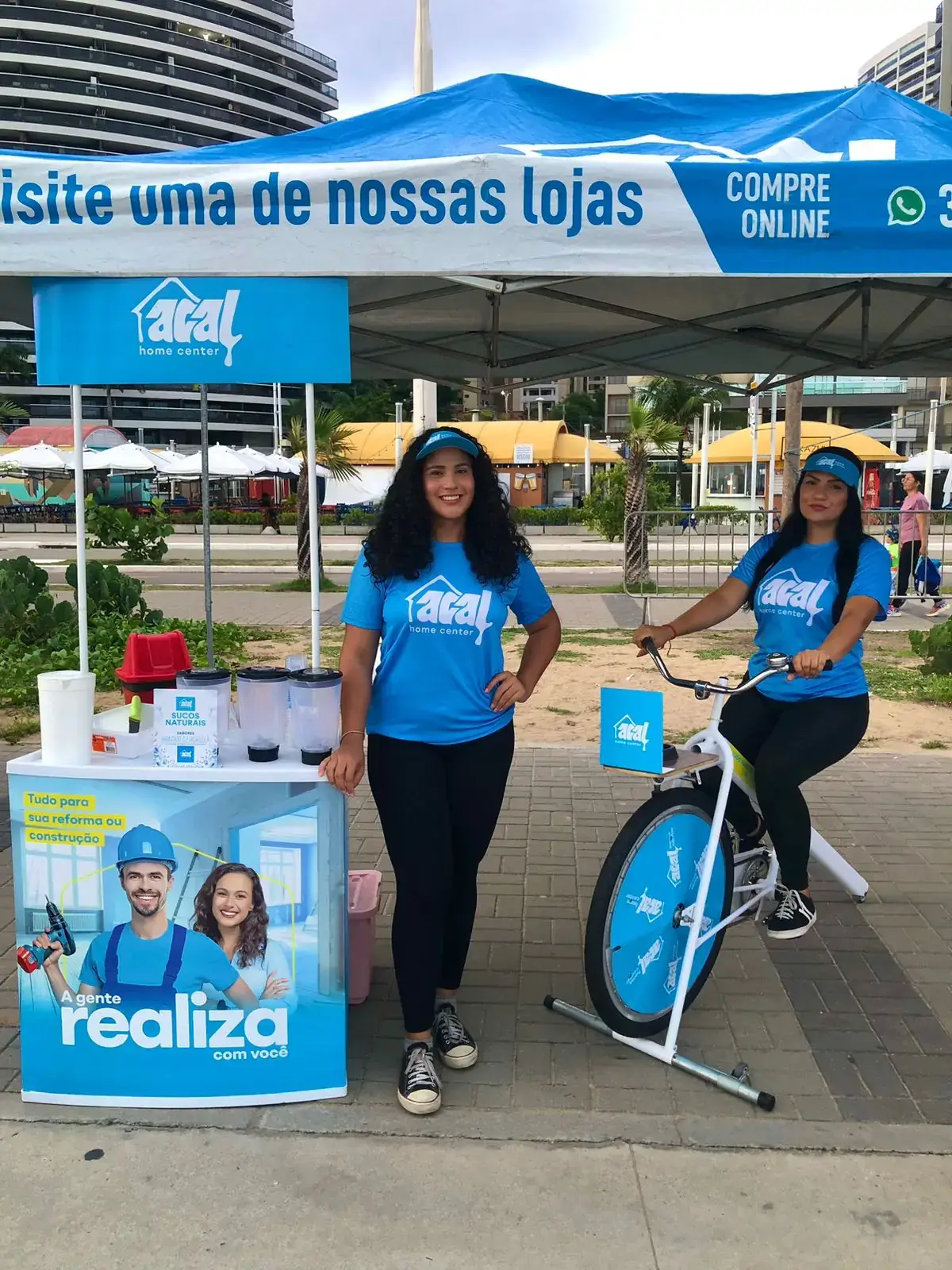 Ação de marketing da Acal home center na orla da Beira-Mar de Fortaleza. Duas promotoras em um estande da marca apresentam uma bicicleta-liquidificador, usada para produzir sucos e interagir com o público.