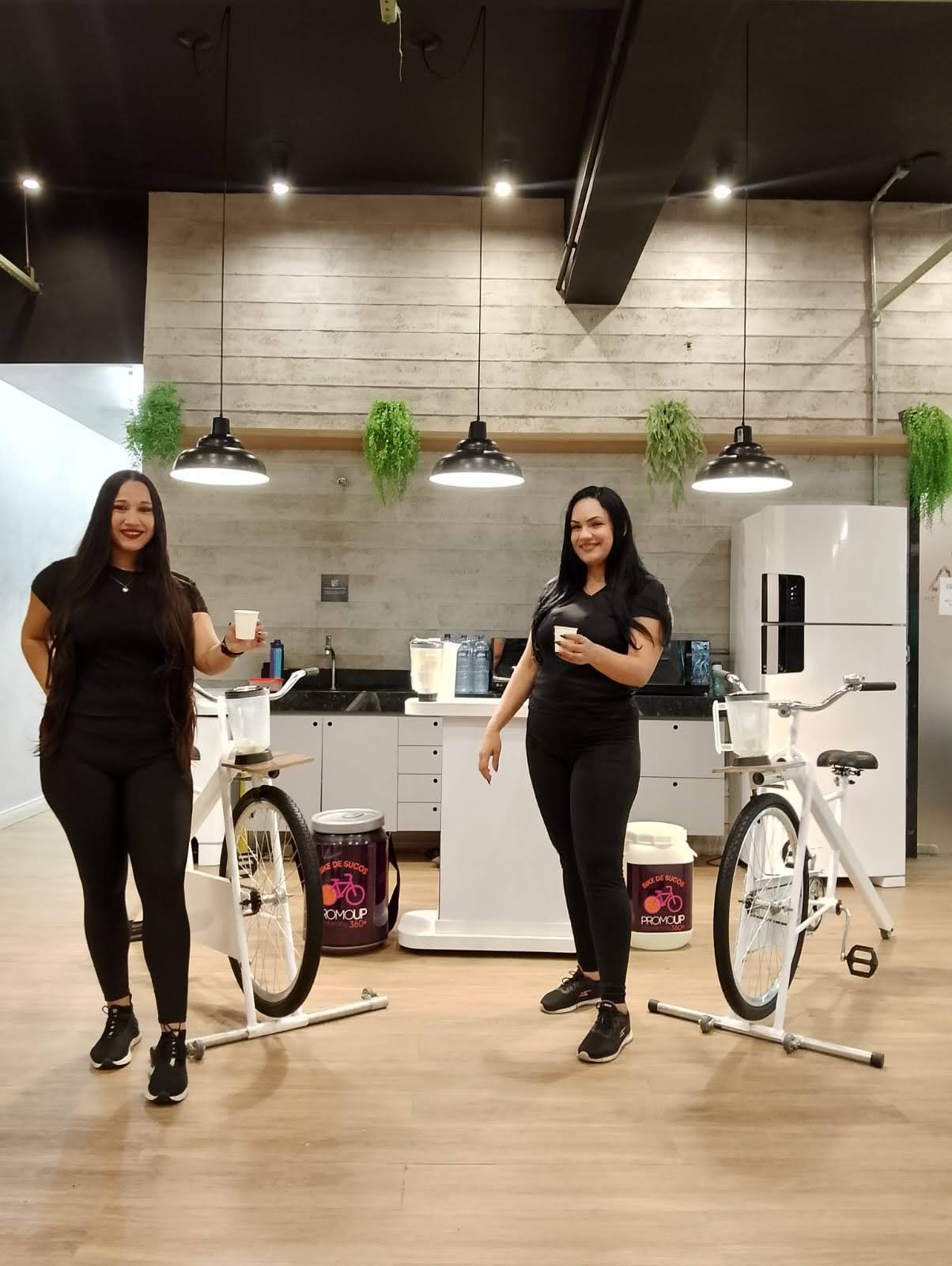 Duas promotoras em ativação de marca demonstram o funcionamento de bicicletas-liquidificador (smoothie bikes). Elas apresentam o resultado da ação: copos com o suco ou smoothie produzido ao pedalar no equipamento.