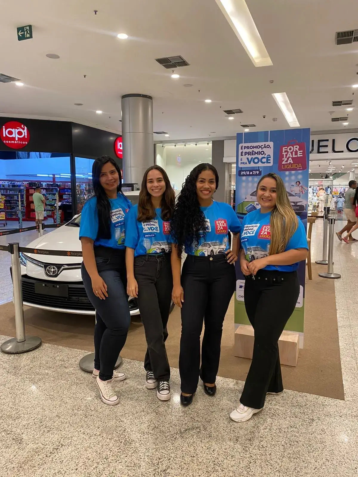 Ação promocional da campanha 'Fortaleza Liquida' em um shopping center. Quatro promotoras de evento, vestindo camisetas azuis da campanha, posam ao lado do carro Toyota branco que está em exposição como prêmio.