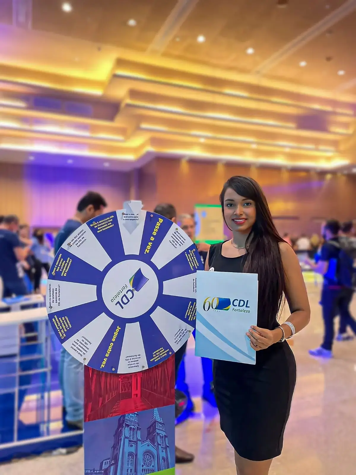 Ação de marketing da CDL Fortaleza em uma convenção ou evento corporativo. Uma promotora apresenta uma roleta de prêmios personalizada, utilizada para interação e engajamento do público, e segura uma pasta com o logo comemorativo dos 60 anos da entidade.