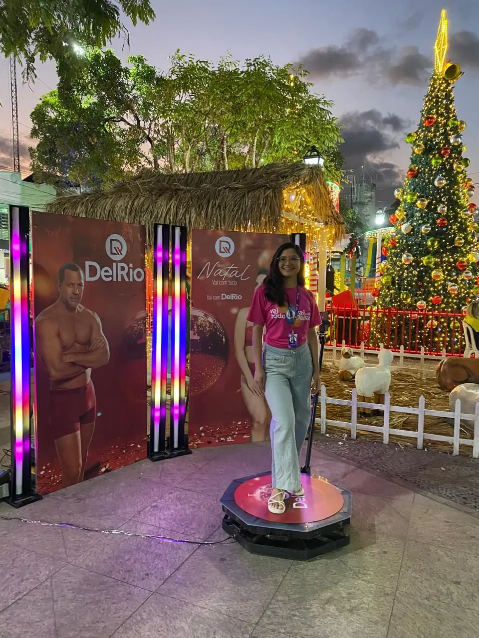 Ação de marketing de Natal da marca DelRio em uma praça pública. Uma participante utiliza uma plataforma de vídeo 360 (spinner) para criar um vídeo interativo, posicionada em frente a banners da campanha e um cenário com árvore de Natal.