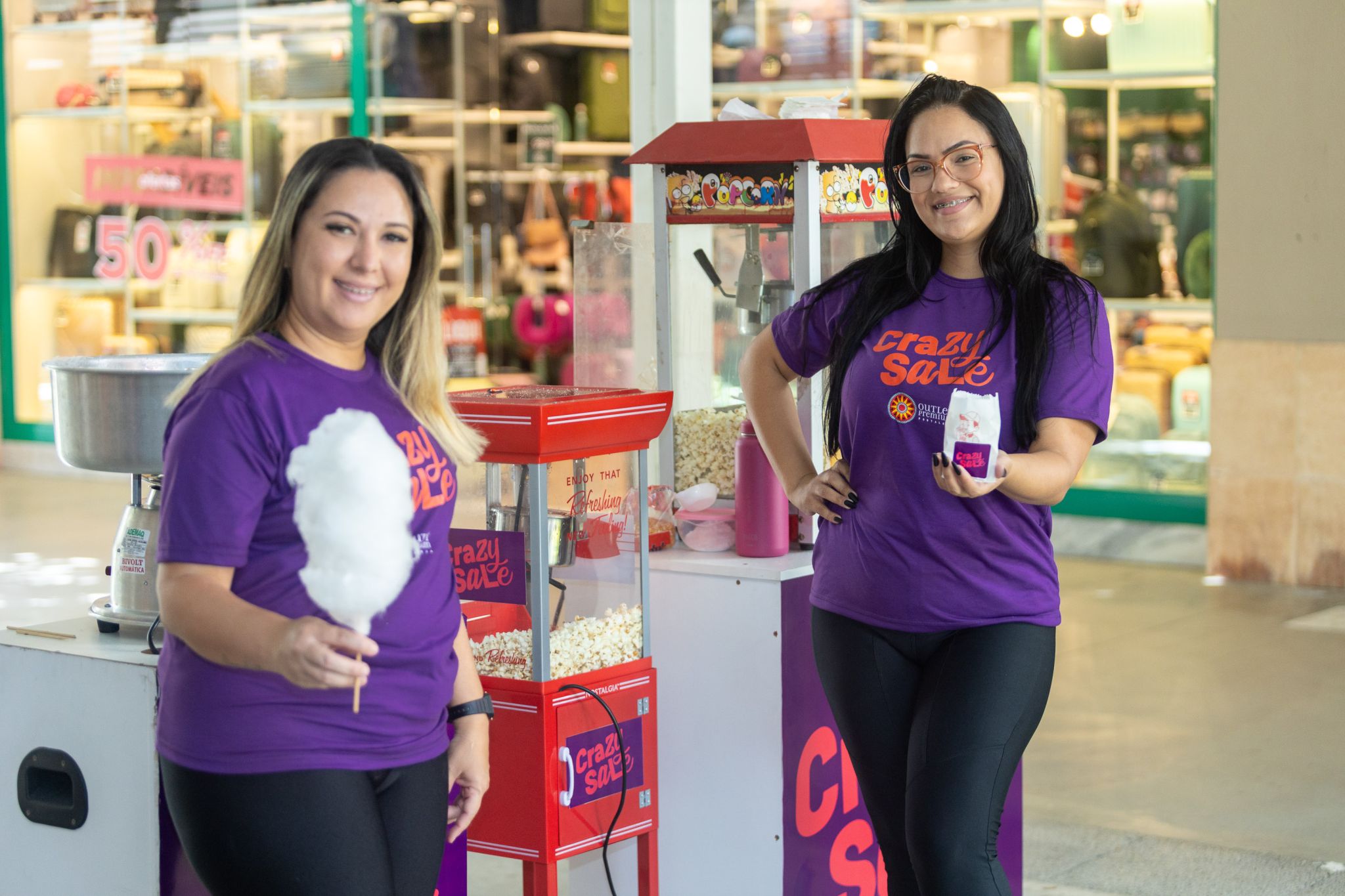 Ação de marketing da campanha 'Crazy Sale' em um shopping center. Duas promotoras uniformizadas distribuem pipoca e algodão-doce como brindes para clientes, ao lado de carrinhos personalizados do evento.