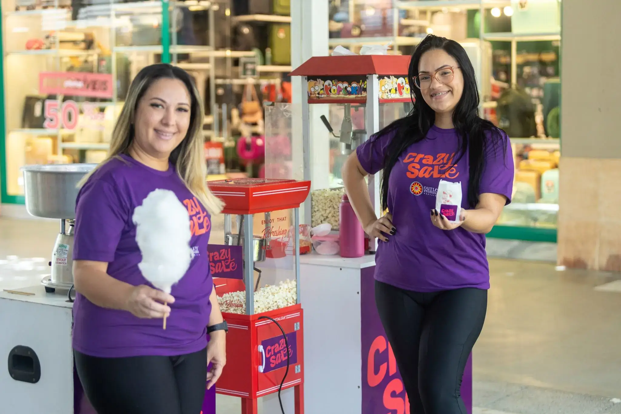 Ação de marketing da campanha 'Crazy Sale' em um shopping center. Duas promotoras uniformizadas distribuem pipoca e algodão-doce como brindes para clientes, ao lado de carrinhos personalizados do evento.