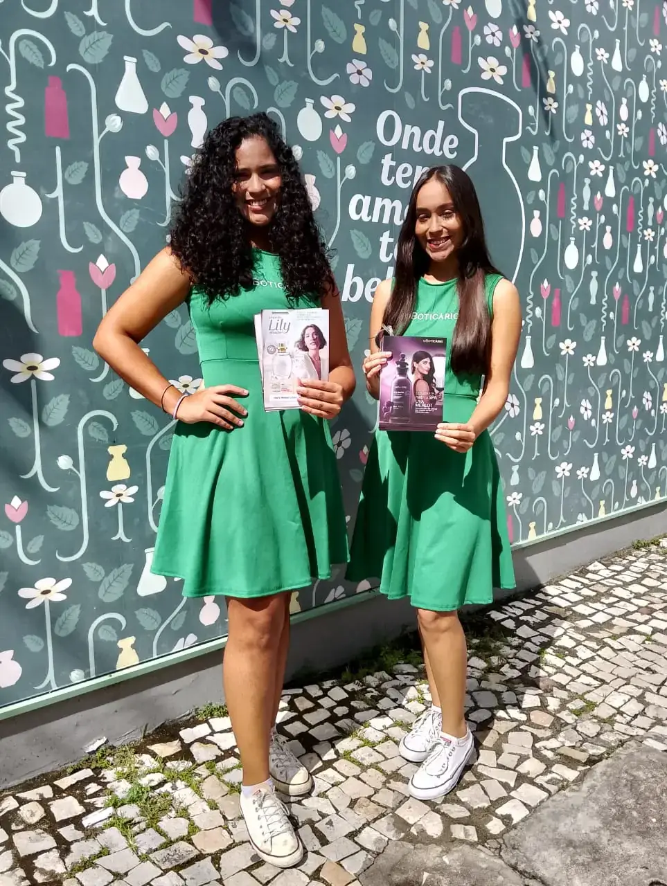 Ação de marketing de O Boticário com duas promotoras divulgando as fragrâncias da marca. Elas vestem uniformes verdes e apresentam folhetos dos perfumes Lily e Malbec em frente a um painel temático da campanha.