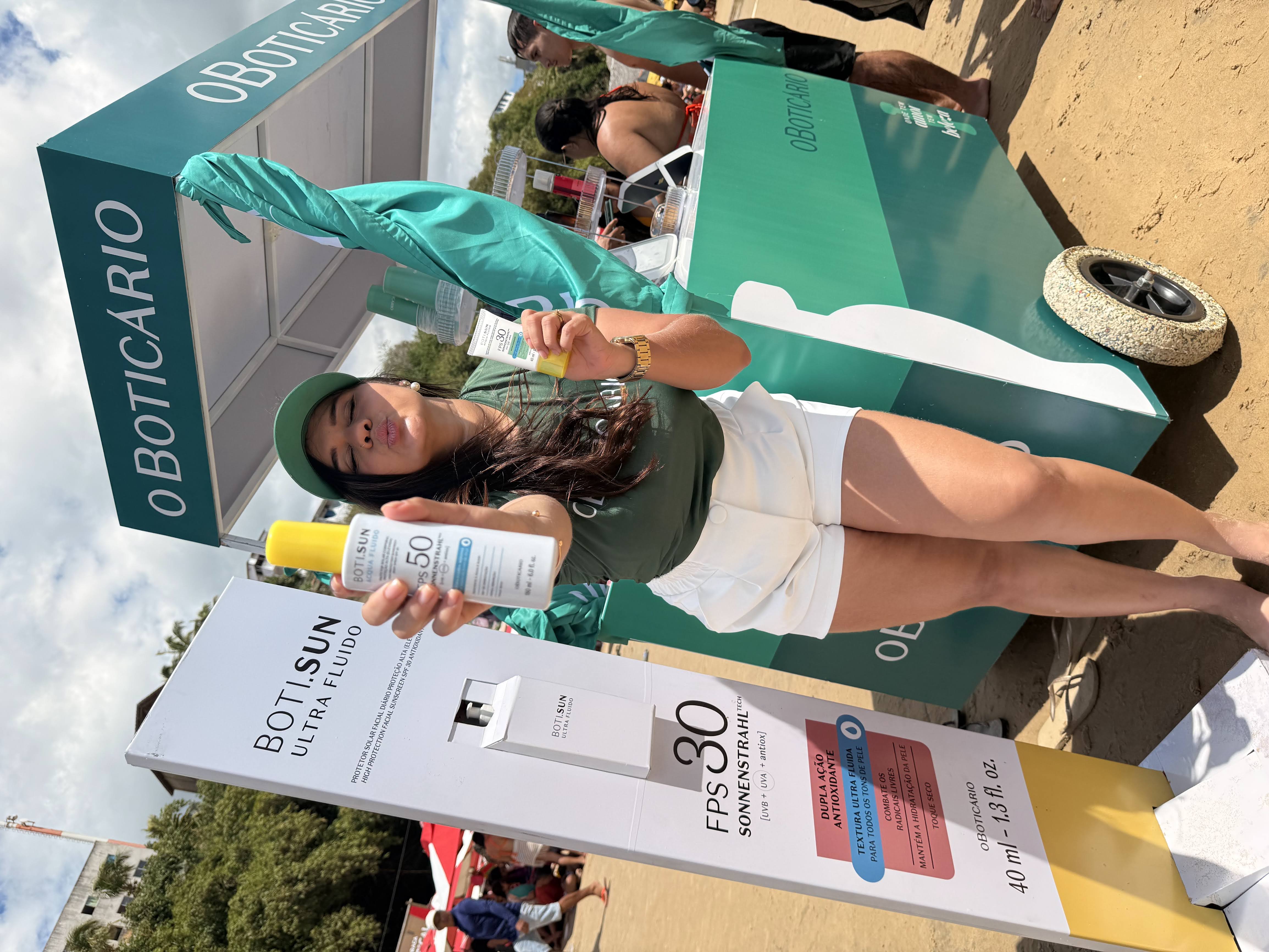 Promotora em ação de marketing de O Boticário na praia, apresentando os protetores solares da linha Boti.Sun. Ela exibe duas embalagens do produto, uma em spray e outra em bisnaga, ao lado de um totem e carrinho personalizados da marca.