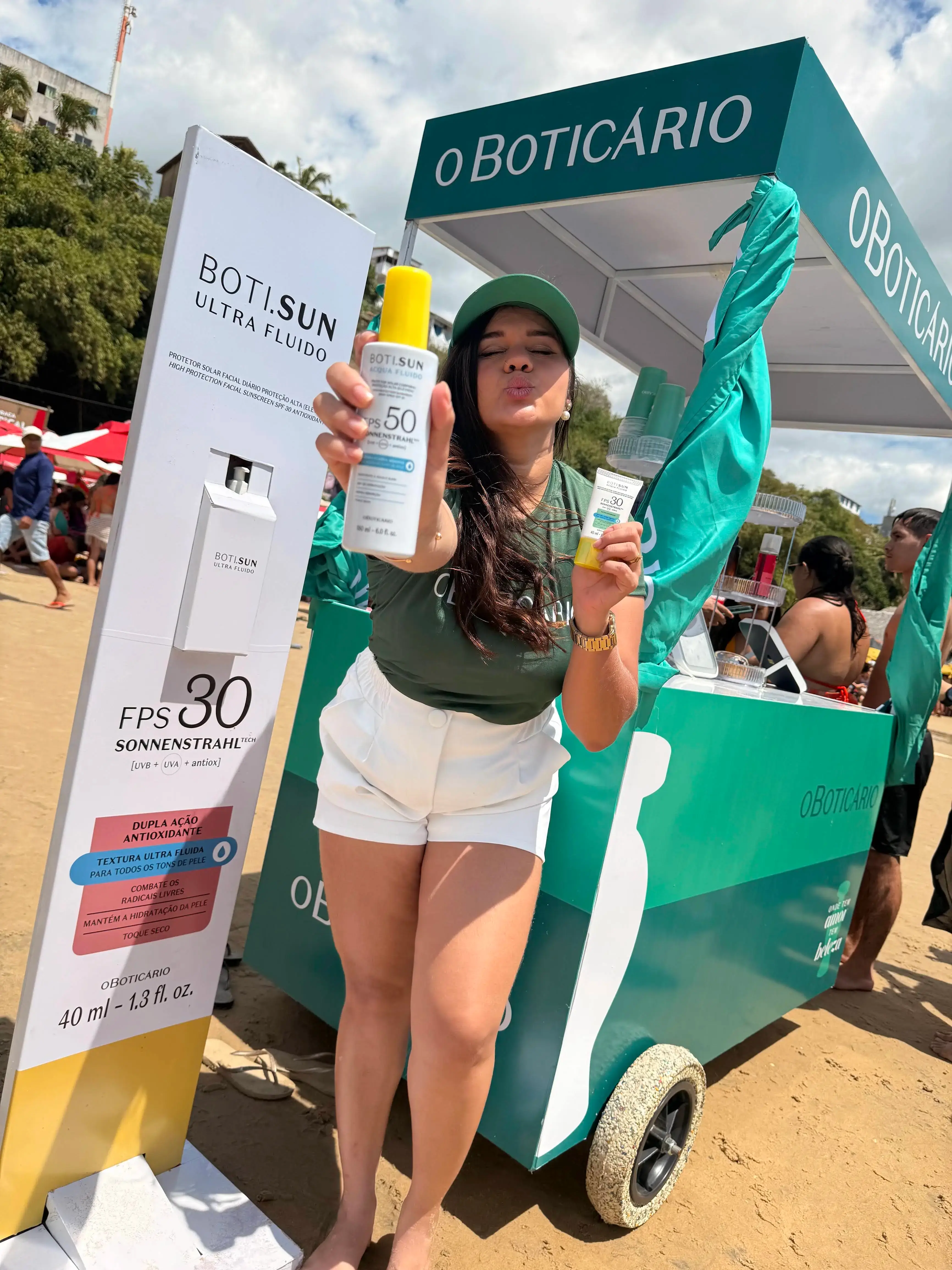 Promotora em ação de marketing de O Boticário na praia, apresentando os protetores solares da linha Boti.Sun. Ela exibe duas embalagens do produto, uma em spray e outra em bisnaga, ao lado de um totem e carrinho personalizados da marca.