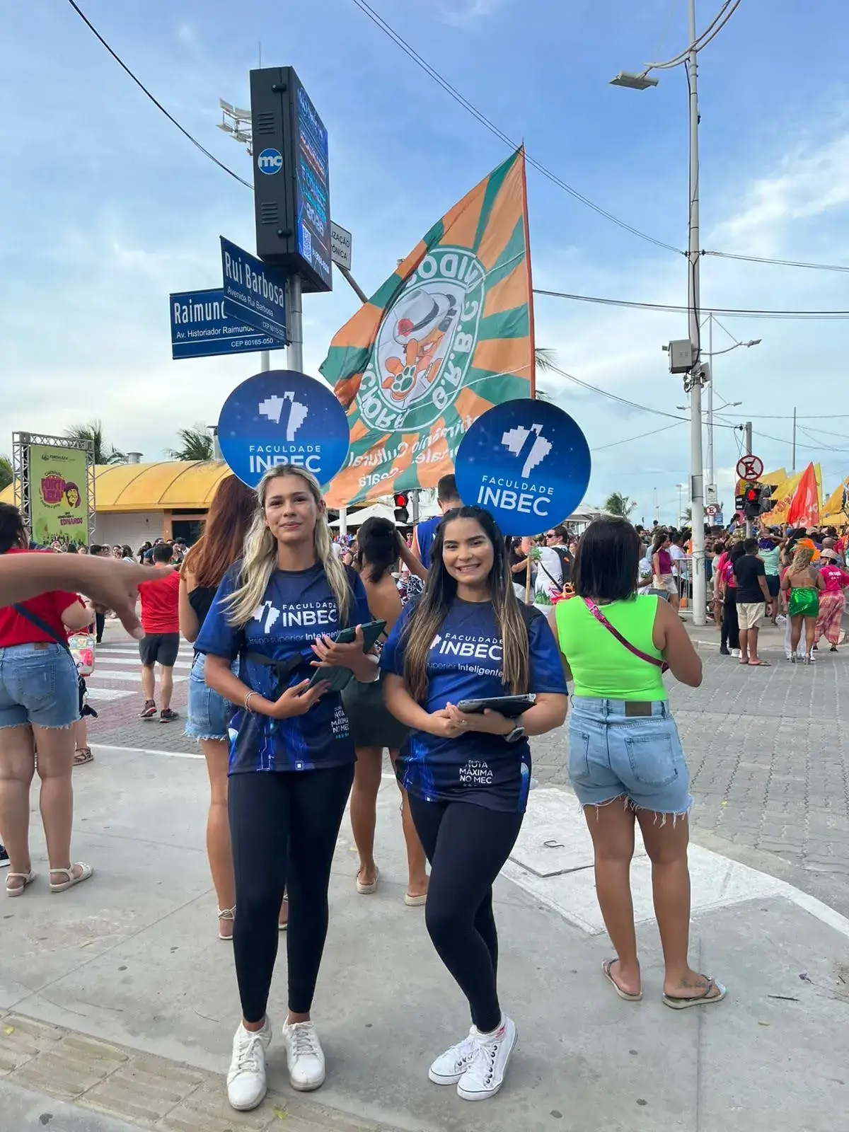 Ação de marketing da Faculdade INBEC com promotoras em um evento de rua movimentado. As duas mulheres vestem uniformes da marca e seguram placas de divulgação (pirulitos) com o logo do INBEC para atrair a atenção e interagir com o público.