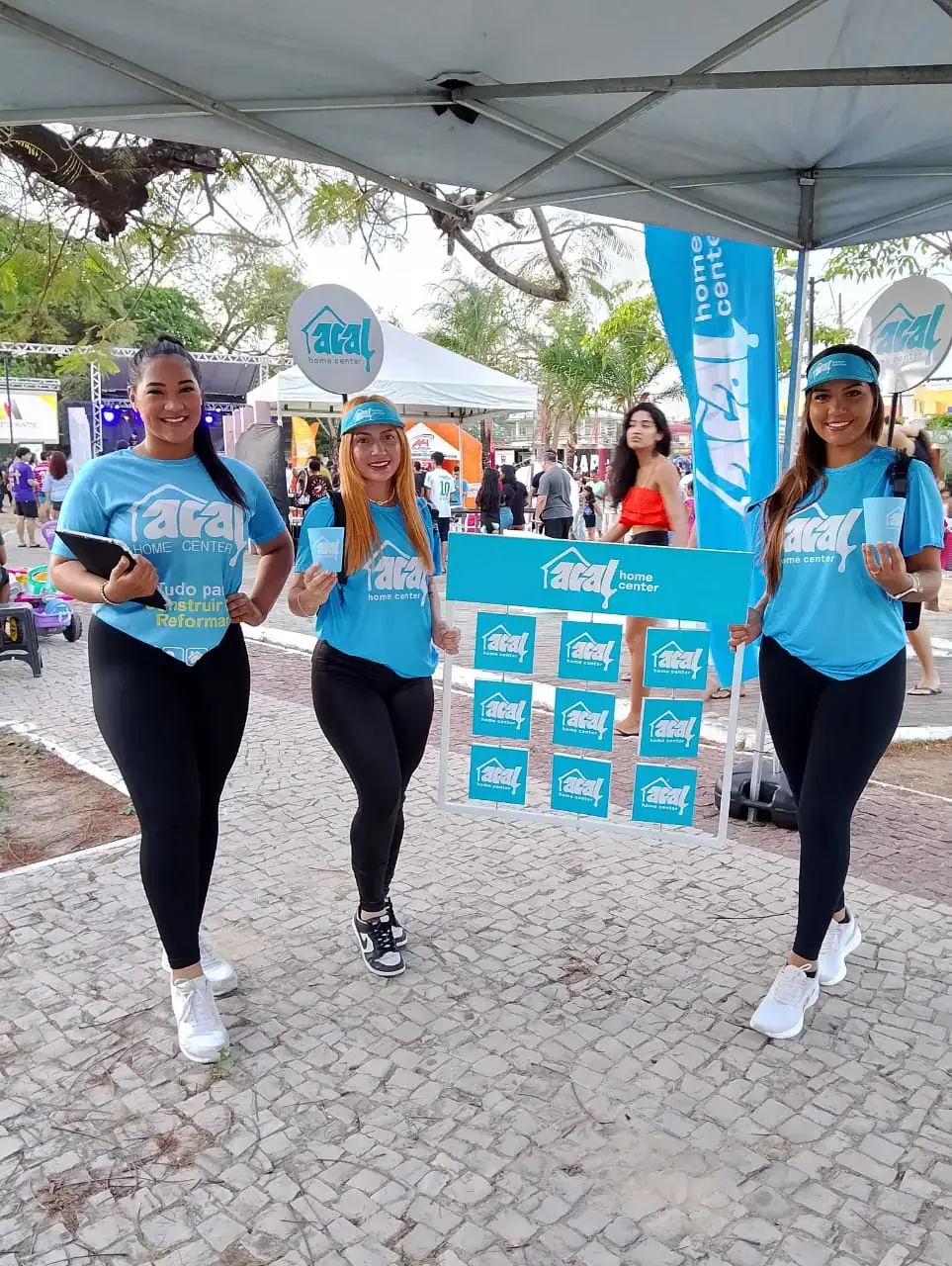Ação de marketing da Acal home center em um evento ao ar livre. Três promotoras uniformizadas interagem com o público, realizando a distribuição de brindes (copos de água) em um estande de divulgação personalizado da marca.