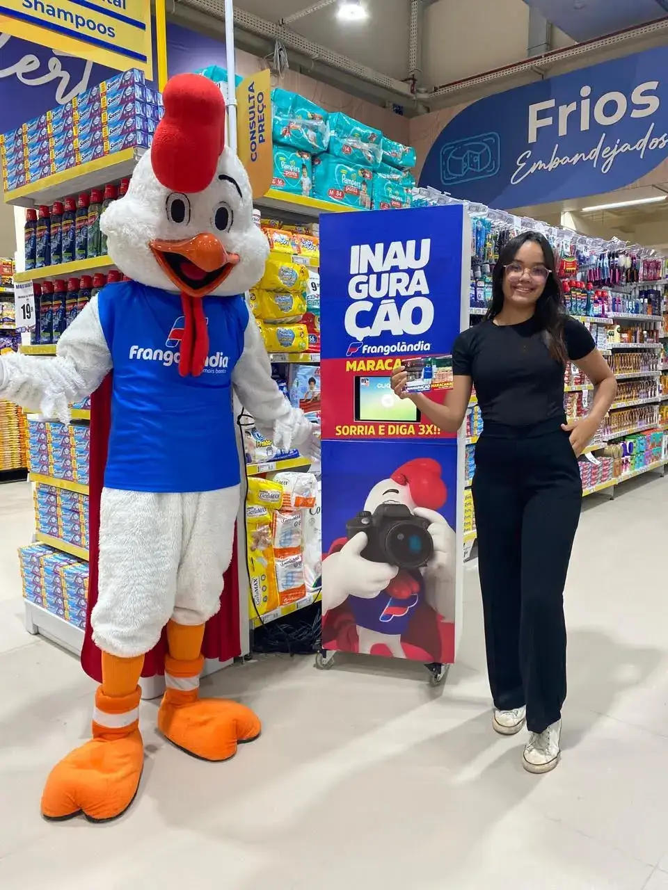Ação de marketing na inauguração do supermercado Frangolândia em Maracanaú. O mascote oficial da marca, um galo, posa para uma foto com uma cliente ao lado de um totem promocional do evento no corredor da loja.