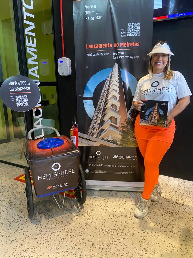 Ação de marketing para o lançamento imobiliário do Hemisphere Residence, da Marquise Incorporações. Uma promotora de eventos apresenta o folder do empreendimento em um estande de divulgação, destacando a localização no bairro Meireles, a 300 metros da Beira-Mar de Fortaleza.